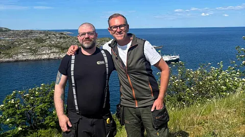 To personer fotografert ved kysten med havutsikt, svaberg og blå himmel i bakgrunnen.